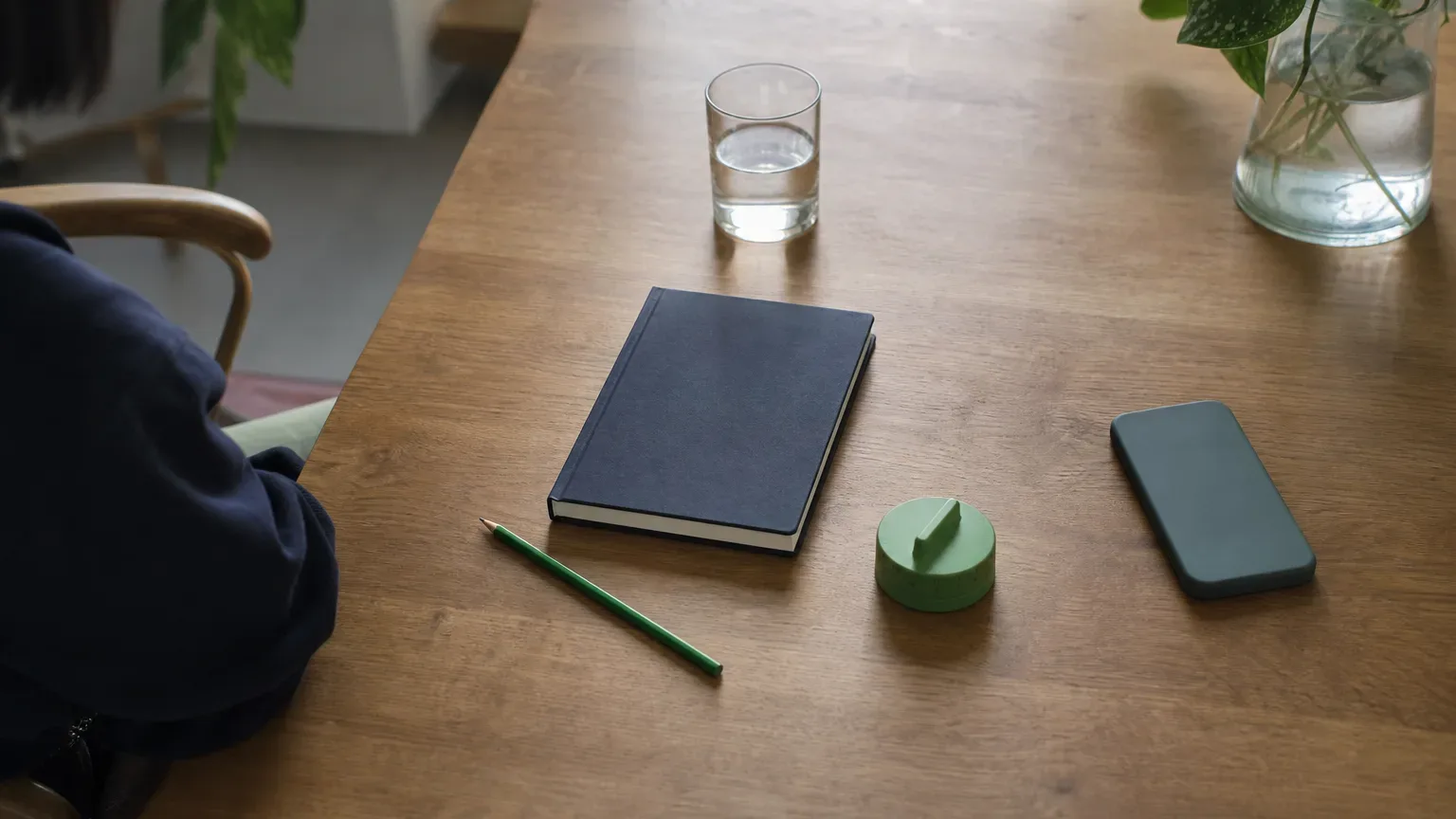 A notebook, pencil, timer, and face-down phone beside a glass of water on a sunlit desk.