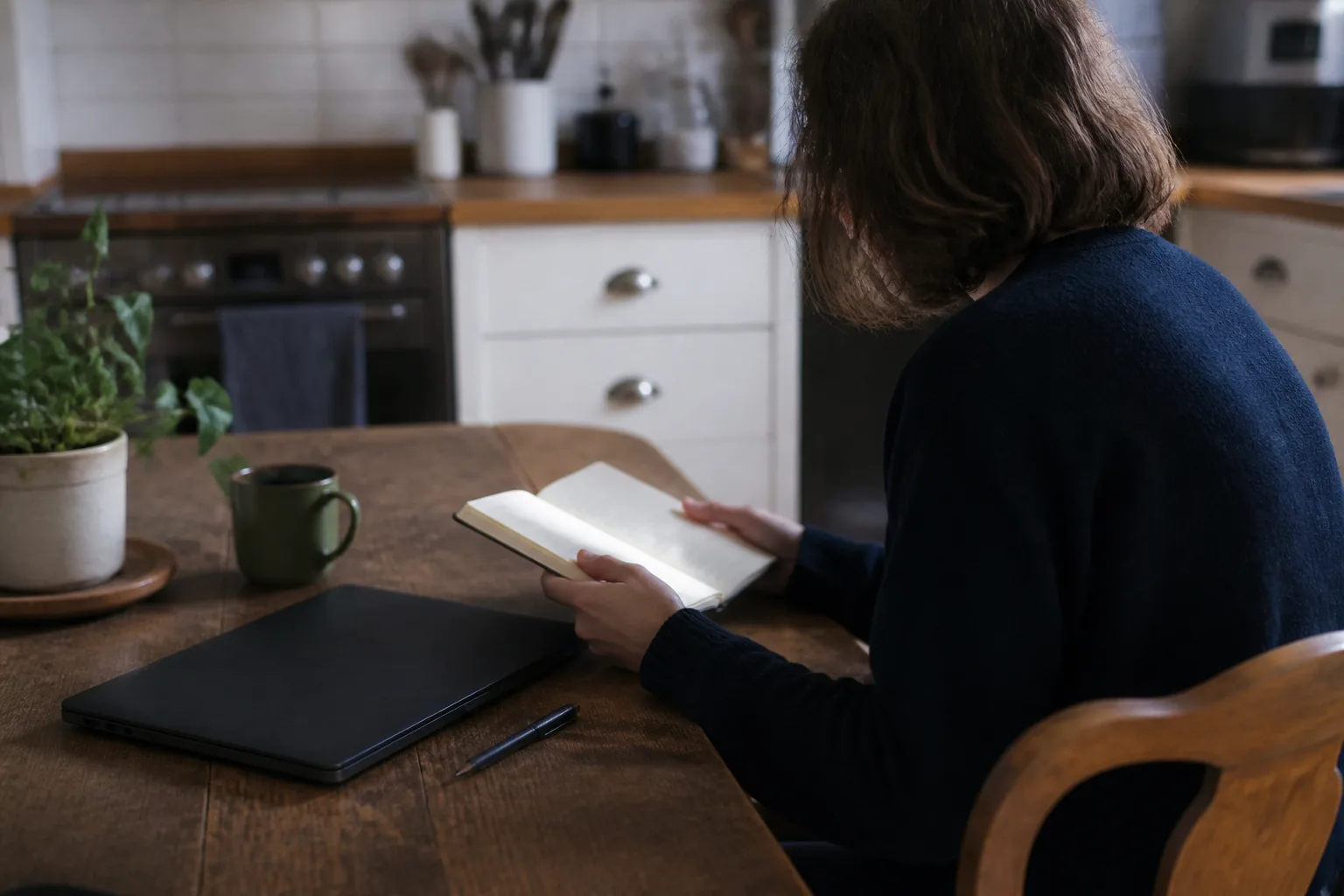 A person pauses over a notebook beside a closed laptop at a kitchen table.